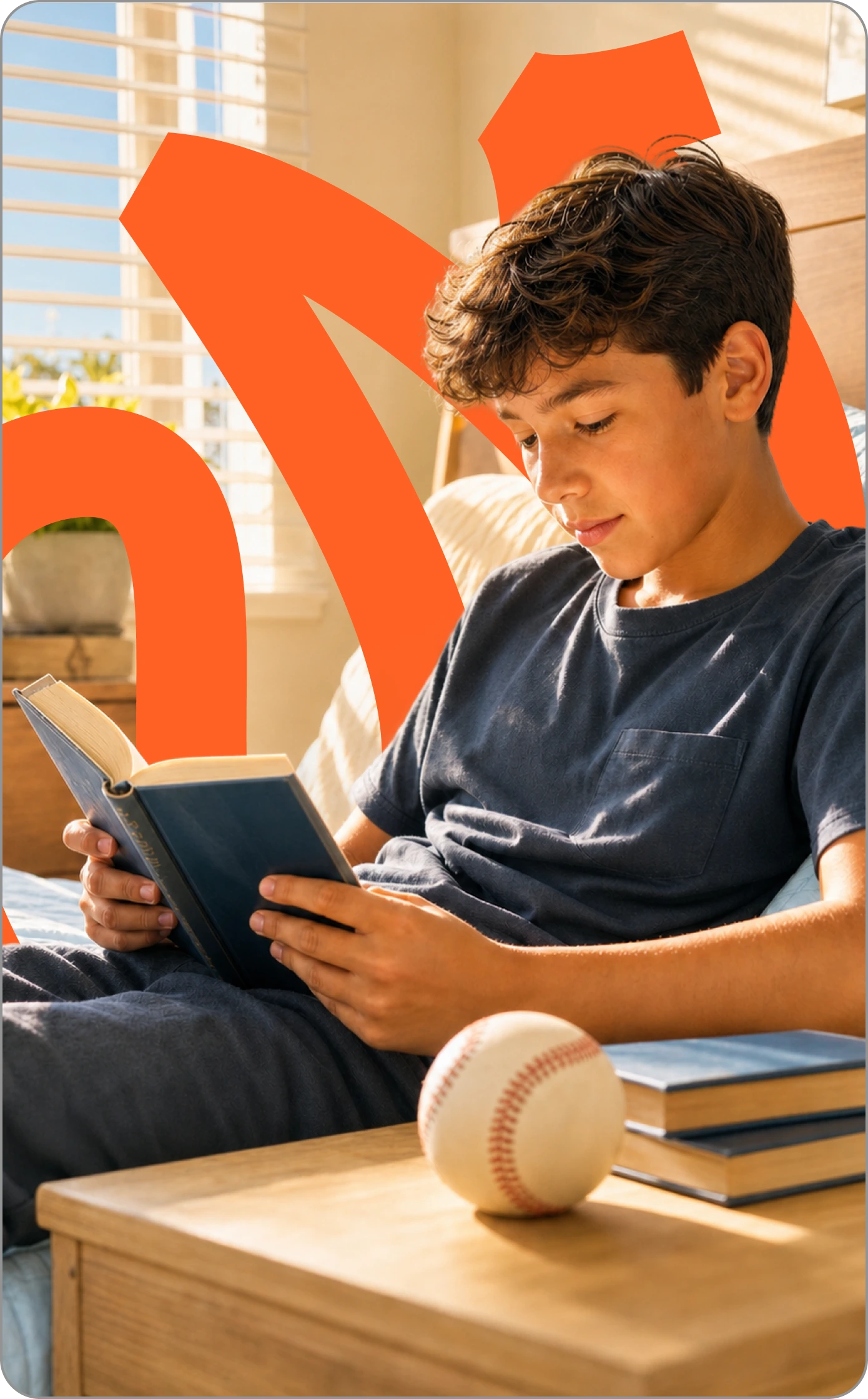 Boy reading indoors with a baseball and books beside him.